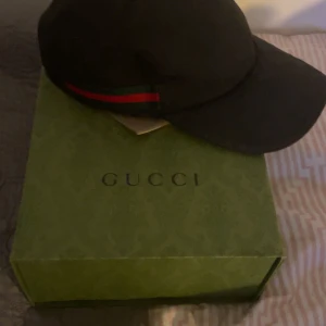 Svart keps från Gucci - Säljer en snygg svart keps från Gucci. Den har den klassiska Gucci-randen i rött och grönt på sidan. Kepsen är i mycket bra skick och kommer med originalförpackning och kvitto. Perfekt för att ge din outfit en lyxig touch! Storlek L.