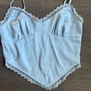 Ljusblå linne med spetsdetaljer - Säljer en söt ljusblå linne med tunna axelband och vacker spetskant. Perfekt för sommardagar eller som ett lager under en kofta. Materialet känns mjukt och luftigt.