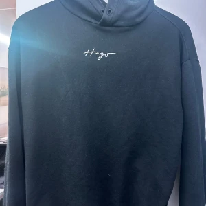 Svart hoodie från Hugo - Säljer en stilren svart hoodie från Hugo med vit broderad logga på bröstet. Perfekt för både vardag och chill. Den har en klassisk passform och är superbekväm. Passar perfekt till jeans eller joggers. Hör av dig om du är intresserad!