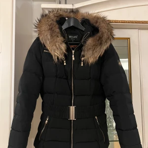 Svart dunjacka från Hollies - Säljer en snygg svart dunjacka från Hollies i storlek 40. Jackan har en fluffig pälsfodrad huva och dragkedja framtill. Den är perfekt för kalla vinterdagar och har en stilren design med guldiga detaljer. Innerfodret har ett coolt leopardmönster. Passar både till vardags och fest!