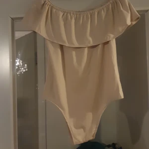 Beige body med volangdetalj - Säljer en superfin beige body med volangdetalj. Den har en off-shoulder design som ger en trendig look. Materialet känns mjukt och bekvämt mot huden. 