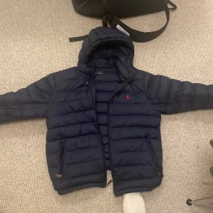 Mörkblå pufferjacka från Polo Ralph Lauren - Säljer en snygg mörkblå pufferjacka från Polo Ralph Lauren i storlek M. Jackan har en klassisk design med dragkedja och det ikoniska röda logotypbroderiet på bröstet. Perfekt för kyliga dagar med sin varma och bekväma passform. 🧥 den säljes på grund av att den är för liten den är M men sitter som S.  Nypris: runt 5000