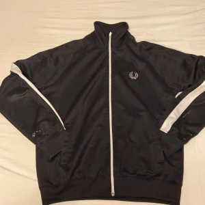 Svart träningsjacka från Fred Perry - Säljer en svart träningsjacka från Fred Perry i bra skick. Jackan har fått lite vit färg längs ärmarna. Perfekt för både träning och vardag. Den har en broderad logga på bröstet och är långärmad. Passar perfekt för höst och vår!