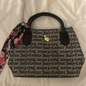 Juicy Couture handväska med blommig scarf - Säljer en snygg Juicy Couture handväska med ett coolt mönster av märkesnamnet i svart på beige bakgrund. Väskan har svarta handtag och en avtagbar axelrem. En söt blommig scarf är knuten på ena handtaget för extra stil. Perfekt för både vardag och fest!
