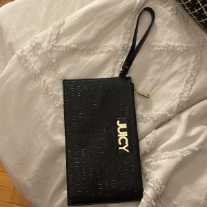 Svart kuvertväska från Juicy Couture - Snygg svart kuvertväska från Juicy Couture med präglad logga och gulddetaljer. Perfekt för en utekväll eller fest! Väskan har en dragkedja och ett praktiskt handtag för enkel bärning. Insidan är fodrad med ett mönster och har flera fack för kort och småsaker.