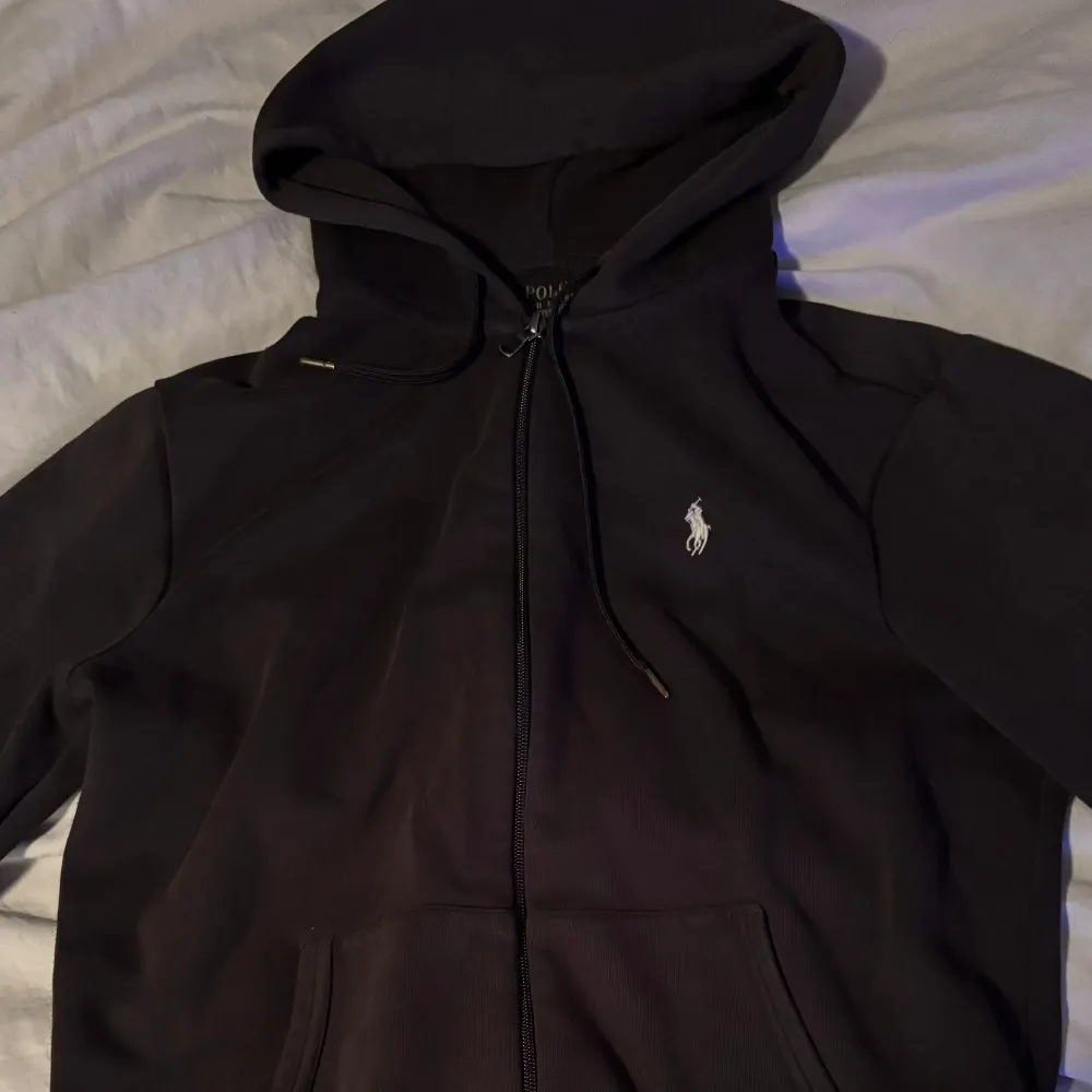 Säljer en stilren svart hoodie från Ralph Lauren med dragkedja och klassisk logga på bröstet. Perfekt för både vardag och chill. Den har en skön passform och är i mycket bra skick. Huvan är justerbar med snören och det finns praktiska fickor framtill.. Tröjor & Koftor.