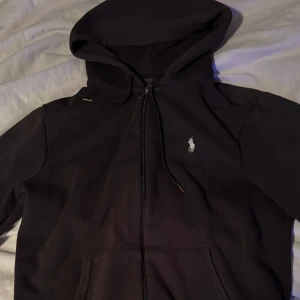 Svart hoodie från Ralph Lauren - Säljer en stilren svart hoodie från Ralph Lauren med dragkedja och klassisk logga på bröstet. Perfekt för både vardag och chill. Den har en skön passform och är i mycket bra skick. Huvan är justerbar med snören och det finns praktiska fickor framtill.