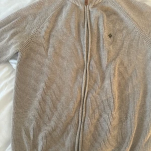 Beige stickad kofta från Morris - Säljer en snygg beige stickad kofta från Morris. Den har en dragkedja framtill och ett diskret broderat emblem på bröstet. Perfekt för höst och vår, med långa ärmar och ribbad krage. Supermysig och stilren!