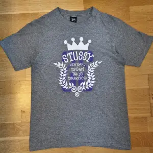 Säljer en grå t-shirt från Stussy i storlek S. Den har ett coolt tryck med texten 'New York, London, Tokyo, Los Angeles' i vitt och lila. T-shirten är gjord av 80% bomull och 20% polyester, vilket gör den både mjuk och hållbar