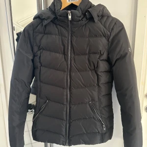 Svart dunjacka med pälsdetalj från Rockandblue - Säljer en snygg svart dunjacka från Rockandblue i mycket bra skick. Jackan har en avtagbar huva med pälsdetalj och dragkedja framtill samt fodrade fickor.Perfekt för kalla vinterdagar! 🖤