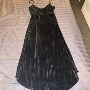 Svart långklänning med smala axelband - Elegant svart långklänning med smala axelband och omlott-design. Klänningen har en vacker plisserad textur som ger en lyxig känsla. Perfekt för festliga tillfällen eller en utekväll. Materialet ser ut att vara mjukt och följsamt, vilket ger en fin passform.