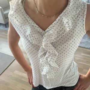 Vit prickig topp - Säljer en söt vit blus med små svarta prickar och volangdetaljer framtill. Blusen är ärmlös och har en v-ringad design som ger en elegant touch. Perfekt för både vardag och festliga tillfällen!
