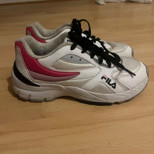 Vita sneakers från Fila - Säljer ett par vita Fila sneakers i bra skick. De har svarta snören och rosa detaljer på sidorna och hälen. Perfekta för vardagsbruk eller träning. Storleken är dam 37. Skorna är bekväma och har en klassisk design med Fila-loggan på sidan.