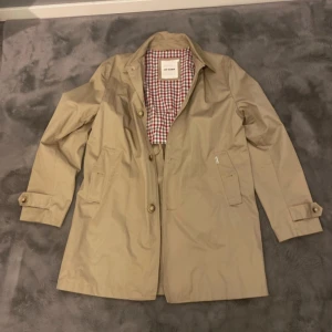 Beige trenchcoat från Ben Sherman - Säljer en stilren beige trenchcoat från Ben Sherman i storlek L. Jackan är som ny och har ett klassiskt rutigt foder i rött och vitt, samt knappar fram och vid ärmsluten. Perfekt för höst och vår! 🧥