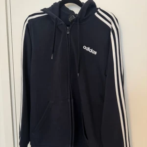 Svart hoodie från Adidas - Säljer en svart hoodie från Adidas i mycket bra skick. Den har de klassiska tre ränderna på ärmarna och en dragkedja framtill. Perfekt för både träning och vardag. Stor logga på bröstet och skön passform. Hör av dig vid intresse!