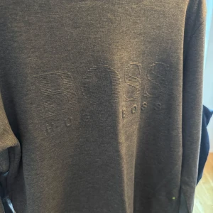 Grå sweatshirt från Boss - Säljer en stilren grå sweatshirt från Boss i mycket bra skick. Tröjan har en diskret broderad logga på framsidan och är gjord i mjukt bomullsmaterial. Perfekt för både vardag och avslappnade tillfällen. Passar till allt!