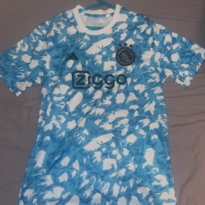 Ajax fotbollströja - Säljer en cool blå och vit fotbollströja från Adidas med ett unikt mönster. Tröjan har korta ärmar och klubbens logga på bröstet. Perfekt för fotbollsfans som vill sticka ut på planen eller läktaren! 🏟️
