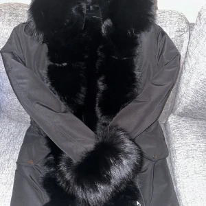 Svart pälsjacka med fuskpälsdetaljer - Säljer en elegant svart jacka med lyxiga pälsdetaljer på krage och ärmslut. Perfekt för kalla vinterdagar när du vill hålla dig varm och stilren. Jackan har en klassisk design som passar både till vardags och festliga tillfällen.