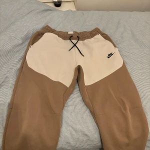 Beige vita Niketech - Säljer ett par snygga och bekväma beige mjukisbyxor från Nike. De har en elastisk midja med snörning och en cool vit detalj på sidorna med Nike-loggan. Perfekta för en avslappnad dag hemma eller en snabb tur till affären. Ett litet slitage som man ser på sista bilden 
