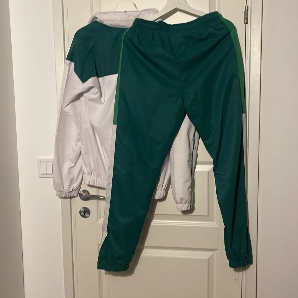 Säljer en snygg vit och grön tracksuit från Lacoste. Jackan har en dragkedja framtill och är långärmad. Perfekt för träning eller en sportig look. Den är i mycket bra skick och har en klassisk stil med Lacostes logga på bröstet.. Jackor.