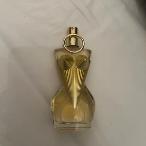 Jean Paul gaultier divine 50ml - Säljer en unik parfymflaska formad som en kvinnokropp. Flaskan är genomskinlig med guldiga detaljer och en elegant design. Perfekt för den som vill ha något annorlunda på sminkbordet. Flaskan har en guldfärgad spraytopp och en dekorativ ringdetalj.