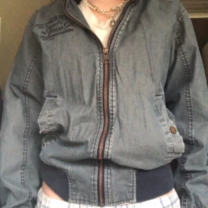 Blå y2k bomberjacka - Säljer en blå bomberjacka med dragkedja och muddar i midjan och ärmsluten. Jackan har knappar på fickorna och en broderad detalj på bröstet. Perfekt för en cool och avslappnad stil.