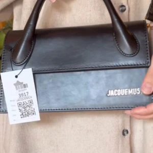 Svart handväska Jacquemus - Snygg svart handväska från Jacquemus med stilren design. Väskan har ett handtag och en elegant logotyp i guld  på framsidan. Perfekt för att ge din outfit en lyxig touch.