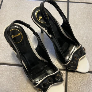 Svarta och vita klackskor från Mossie - Snygga klackskor från Mossie i svart och vitt med dekorativa stenar framtill. Skorna har en öppen tå och en elegant klack, perfekt för att lyfta din stil. De har en stilren design med en touch av glamour.