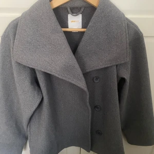 Grå kappa från Gina Tricot - Snygg grå kappa från Gina Tricot med stora knappar och bred krage. Perfekt för höst och vår. Jackan har en stilren design och är lätt att matcha med olika outfits.