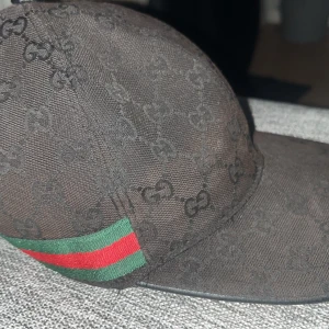 Svart keps från Gucci - Snygg svart keps från Gucci med det ikoniska GG-mönstret. Kepsen har en grön och röd randdetalj på sidan som ger en lyxig touch. Perfekt för att lyfta din stil till nästa nivå.