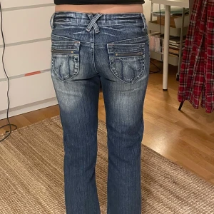Blå jeans med broderade bakfickor - Snygga blå jeans med broderade detaljer på bakfickorna och en unik korsad bältesdesign. De har en klassisk bootcut-stil och är perfekta för en avslappnad look. Jeansen har en knapp och dragkedja framtill. Innerbenslängden:71 cm midja:36cm Jag tar inte emot retur ❤️