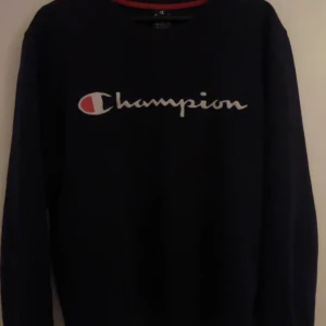 Svart sweatshirt från Champion - Säljer en svart sweatshirt från Champion med deras ikoniska logga broderad på bröstet. Tröjan har långa ärmar och en rund halsringning med röd detalj på insidan. Perfekt för en avslappnad stil.