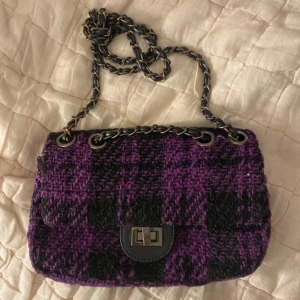 Rutig väska - Snygg axelväska i lila och svart rutigt mönster med kedjeaxelrem. Väskan har en praktisk dragkedja inuti och ett stilrent spänne framtill. Försvunnit lite färg från det silvriga se bilderna!