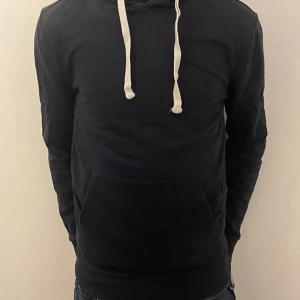 Mörkblå hoodie med vita snören - Säljer en stilren mörkblå hoodie från New Look. Den har en klassisk design med vita snören och en praktisk magficka. Perfekt för en avslappnad stil. Skriv om ni har funderingar ch priset är inte hugget i sten!
