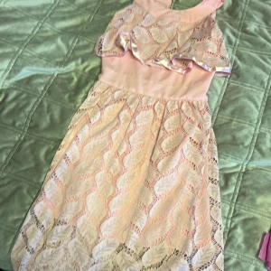 Rosa spetsklänning med volangdetaljer - Charmig rosa klänning i spets med volangdetaljer över axlarna. Klänningen har en elegant omlott-design och är ärmlös, vilket gör den perfekt för varmare dagar. Den har en mjuk rosa nyans och ett vackert mönster som ger en romantisk känsla.🩷💞