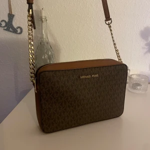 Brun axelväska från Michael Kors - Snygg brun axelväska från Michael Kors med guldfärgade kedjedetaljer och justerbar axelrem. Väskan har ett klassiskt MK-mönster och en dragkedja upptill. Använd max 3 gånger så i nyskick!