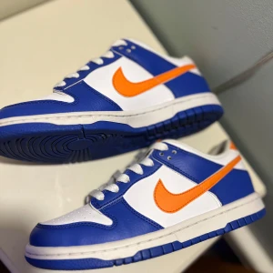 Nike Dunk i blått och vitt med orange detaljer - Snygga Nike Dunk sneakers i en fräsch färgkombination av blått och vitt med en iögonfallande orange swoosh. Skorna har klassisk snörning och en bekväm passform, perfekt för en sportig look.