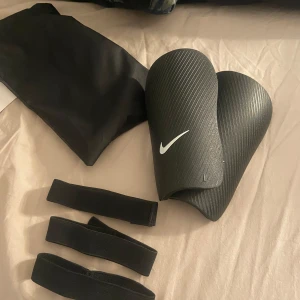 Nike J Guard benskydd - Säljer ett par svarta Nike J Guard benskydd för fotboll. De är lätta och smidiga med en vit Nike-logga. Inkluderar elastiska remmar för säker passform och en praktisk förvaringspåse. Helt oanvända