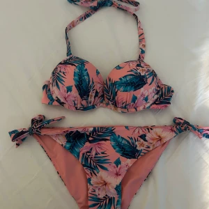 Blommig bikini med halterneck knytband - Snygg bikini i rosa med tropiskt blommönster i blått och grönt. Överdelen är push up, osäker på storlek då lapparna inte är kvar men skulle säg att det är en A-kupa och att storleken på underdelen är S/M.