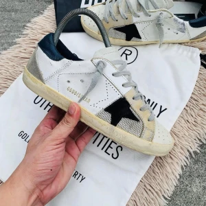 Golden Goose sneakers med stjärndetalj - Snygga Golden Goose sneakers i vitt med en svart stjärndetalj på sidan. Skorna har grå snörning och en kontrasterande mörkblå häl. Tillverkade i skinn och mocka för en stilren look.