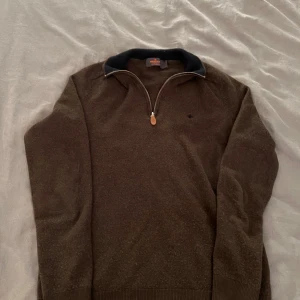 Brun tröja med dragkedja från Morel - Säljer en brun tröja från Morel med dragkedja och mörkblå krage. Tröjan har ett litet broderat emblem på bröstet och är tillverkad i 100% ren ull. Perfekt för kyliga dagar!