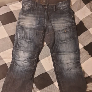 Blå cargobyxor från G-Star - Snygga blå cargobyxor från G-Star med flera praktiska fickor och knappar. Byxorna har en cool tvättad look och är perfekta för en avslappnad stil. De har en normal passform och är tillverkade i slitstarkt material.