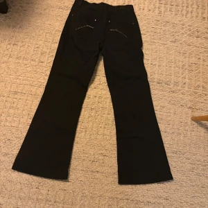 lågmidjade jeans  - Snygga svarta byxor från Olsen med bootcut-stil. Byxorna har dekorativa detaljer med nitar på bakfickorna som ger en extra touch. Perfekta för en stilren look.