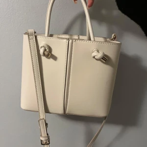 Beige handväska ZARA - Beige handväska med avtagbar axelrem. Väskan har en dragkedja och magnetknappar. Ursprungligen från Zara. Använd fåtal gånger. Ser ut som ny.