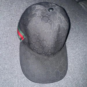 Svart keps från Gucci - Snygg svart keps från Gucci med det klassiska GG-mönstret. Kepsen har en grön och röd randdetalj på sidan och justerbar rem baktill. Perfekt för att ge en lyxig touch till din outfit.