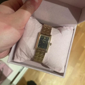 Guldfärgad klocka med svart urtavla - Elegant guldfärgad klocka från Himo med en rektangulär boett och svart urtavla. Klockan har romerska siffror och en länkarmband i metall. Perfekt för en stilren look. Samt vattentåligt 