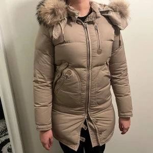 Beige dunjacka från Cedrico - Säljer en snygg beige dunjacka från Cedrico med pälskantad huva. Jackan har dragkedja och knappar framtill samt praktiska fickor med dragkedja. Perfekt för kalla vinterdagar.