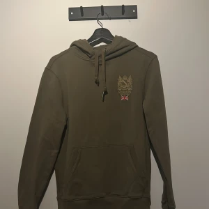 Olivgrön/Beige hoodie från Morris - Säljer en snygg olivgrön/Beige hoodie från Morris med broderad logga och Union Jack-detalj på bröstet. Tröjan har en klassisk känguruficka och justerbar huva med snören. Perfekt för en avslappnad stil.