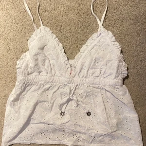 Vit broderad linnetopp från Censored - Söt vit linnetopp från Censored med broderade detaljer och smala axelband. Toppen har en v-ringning och en knytning framtill. 