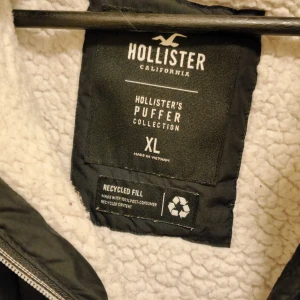 Svart pufferjacka från Hollister - Säljer en svart pufferjacka från Hollister med dragkedja och fuskpälsdetaljer på huvan. Jackan har långa ärmar och två fickor med knappar framtill. Perfekt för kyliga dagar.